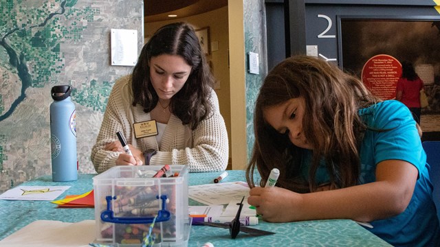 Two girls, one slightly older and wearing a name tag, sit at a table with art supplies using markers