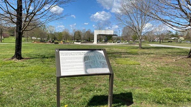 A wayside exhibit in a city park with a large bronze bell in the distance.