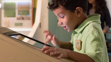 A kid in a green shirt touches a screen with an excited face.