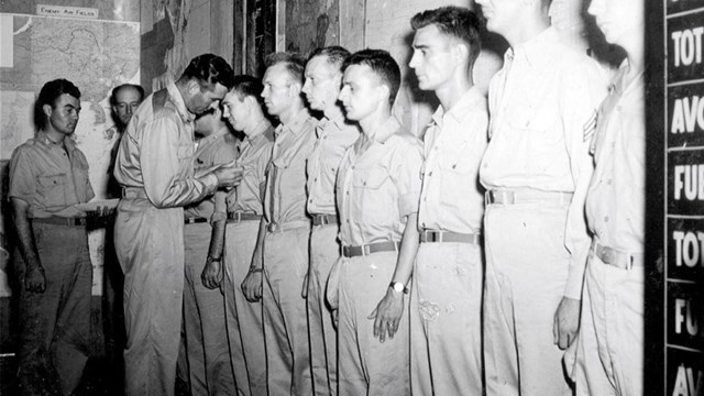 Men stand in a line as a man pins something on the shirt of one of the men.