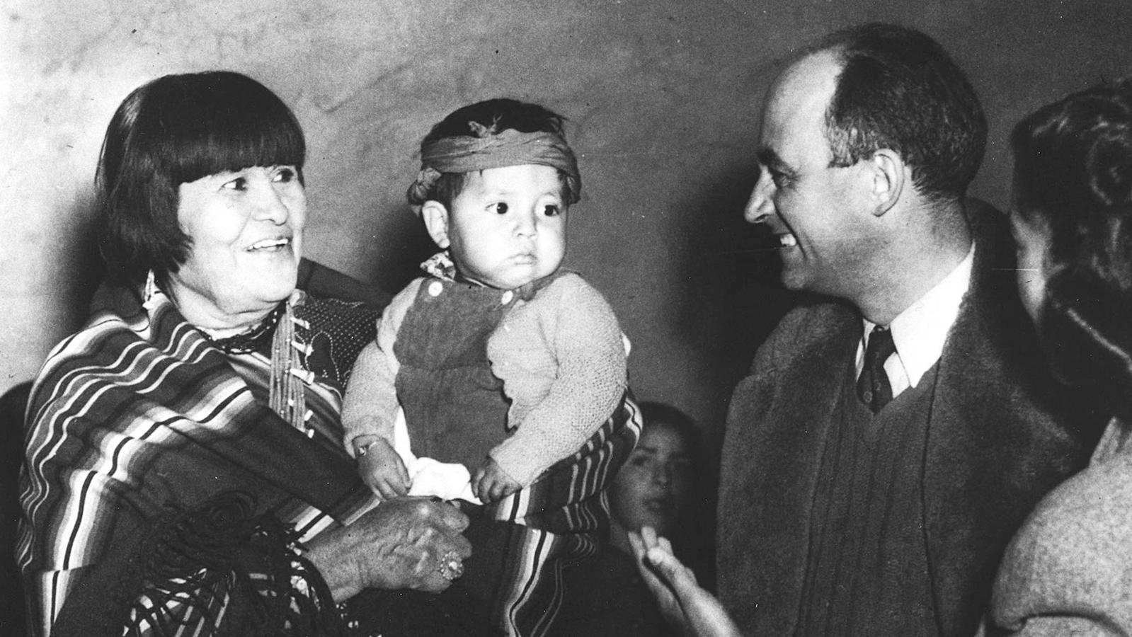 A black and white image of a lady with short black hair holding a baby in front of a couple.&nbsp;