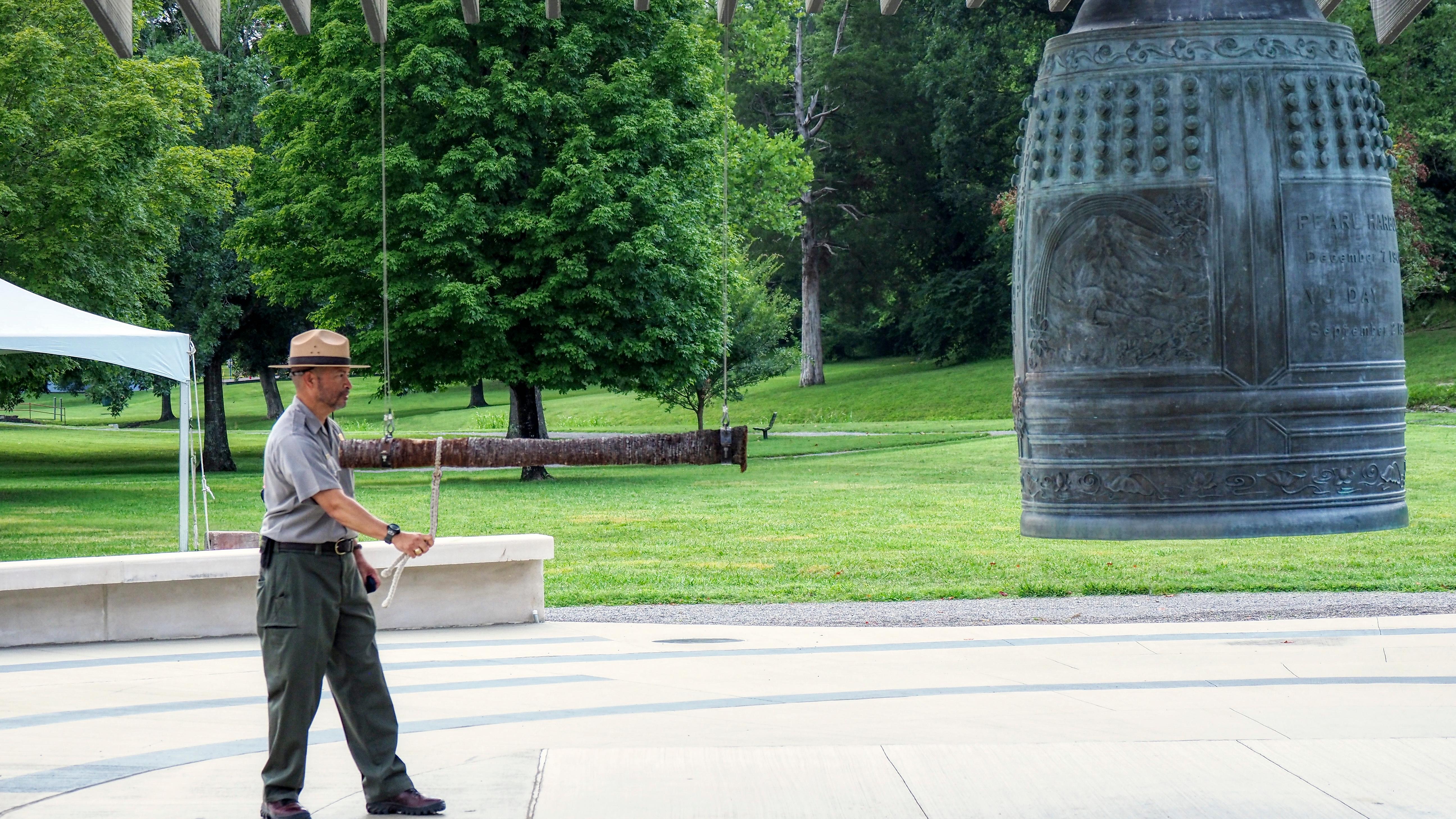 Park ranger faces a large bronze bell overhanging a courtyard about to be rung by a wooden striker.