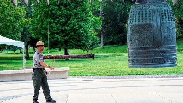 Park ranger faces a large bronze bell overhanging a courtyard about to be rung by a wooden striker.