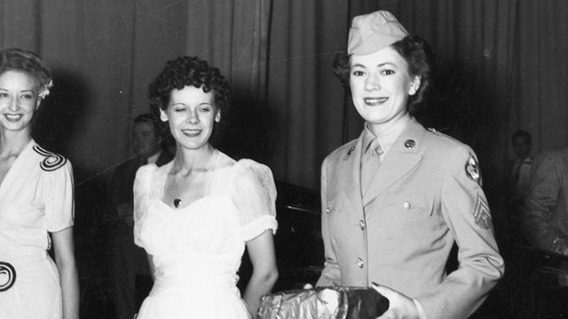 A black and white photo of a woman in uniform holding a package standing beside women in ball gowns.