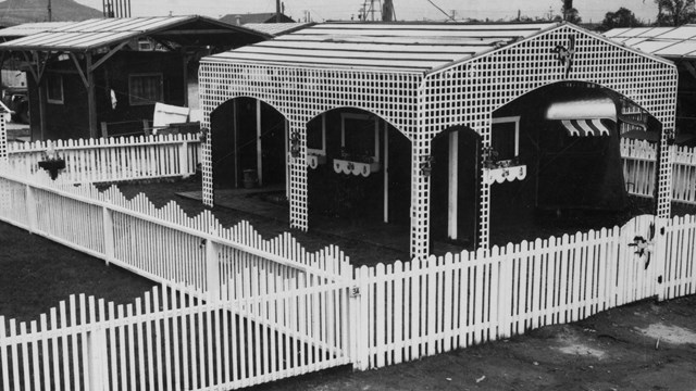 Black and white photo of a small dwelling with a white picket fence. 