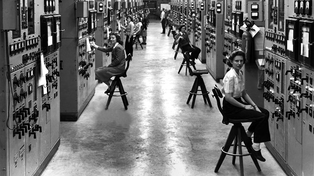 A black and white photo of women sitting on chairs on either side of an aisle. 