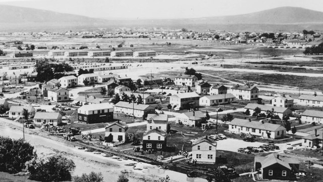 Black and white photo of houses along a river with two large hills in the distance. 