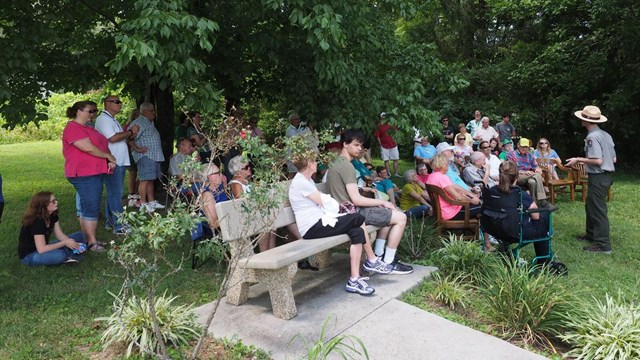  A man in a ranger uniform speaks to a group of about 30 seated people in a park 