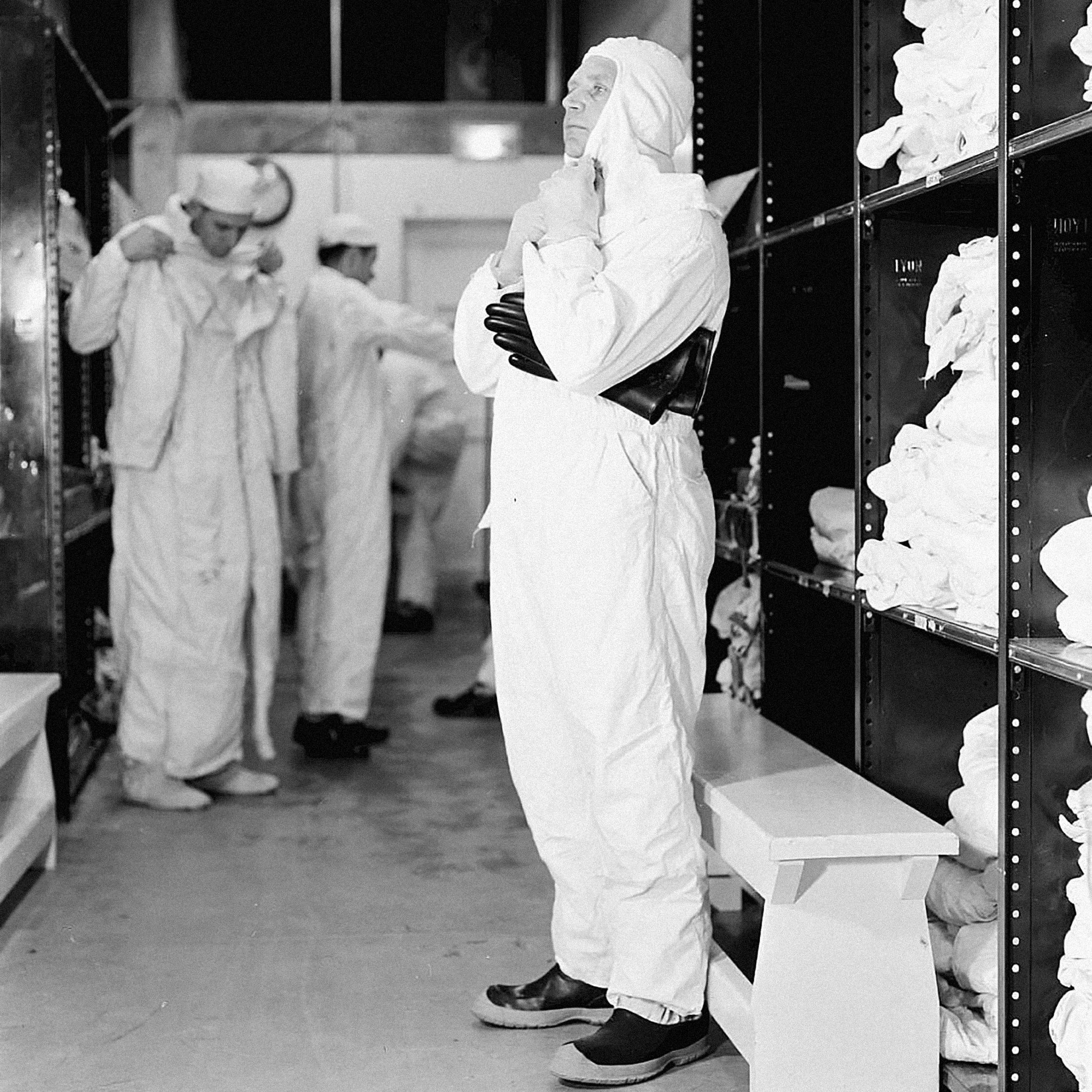 A man suits up in a white coverall suit with other men doing the same in the background.
