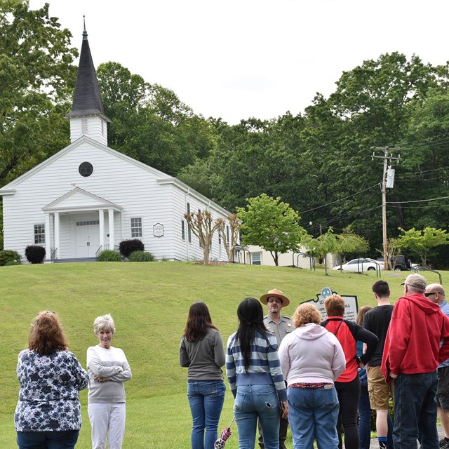 People gather on a hillside in front of a white structure with a steeple.