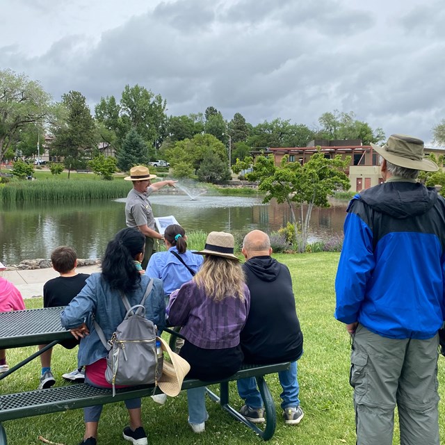 Visitors stand in a grassy area facing a ranger in uniform as he points at the lake behind him.