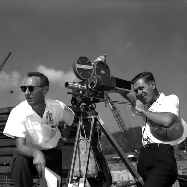 A black and white image of two guys looking through a large camera.