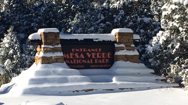 Dark brown sign with light-red letters, hangs between 2 snow covered native sandstone pillars 