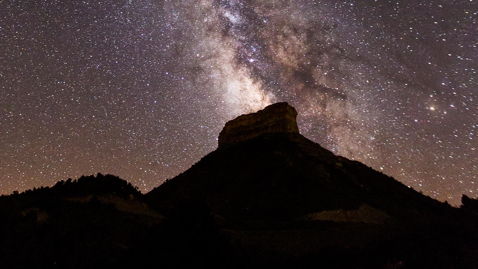 A starry sky bisected by the Milky Way rises above the prominent mesa of Point Lookout.