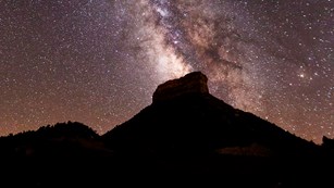 A starry sky bisected by the Milky Way rises above the prominent mesa of Point Lookout.