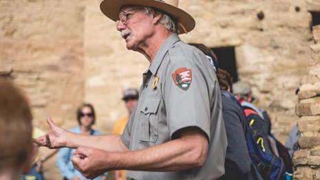A ranger with white hair and mustache gestures with hands while visitors gather around listening