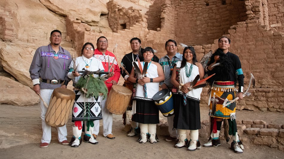 Eight Pueblo dances dressed in regalia stand in the plaza in front of an ancestral site