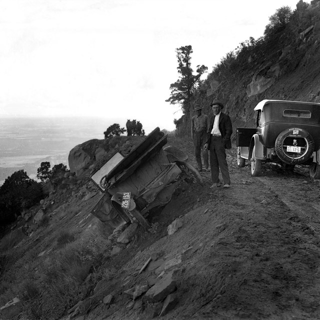 Two early vehicles on a dirt road that traverses a steep hillside. One is has gone off the road.