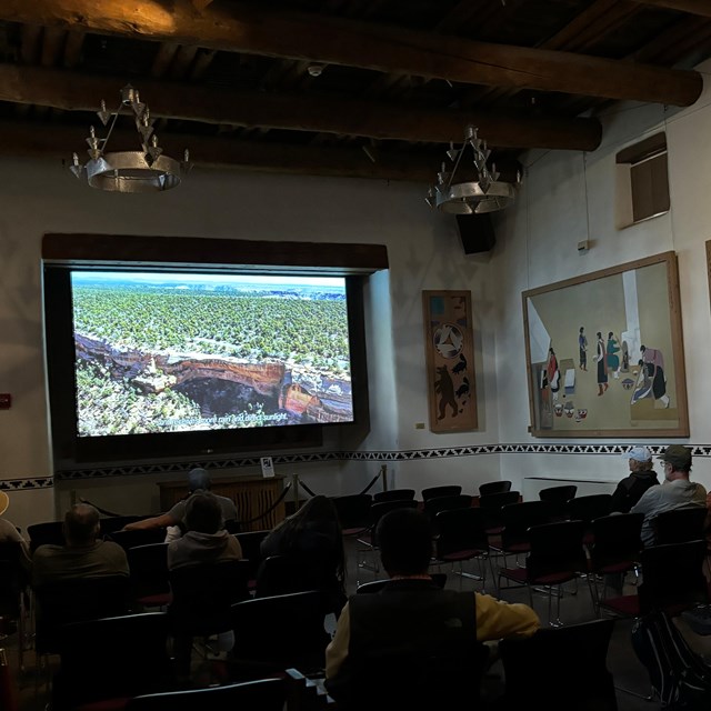 Visitors watch the park video in a dimly lit room that highlights Indigenous wall  murals. 