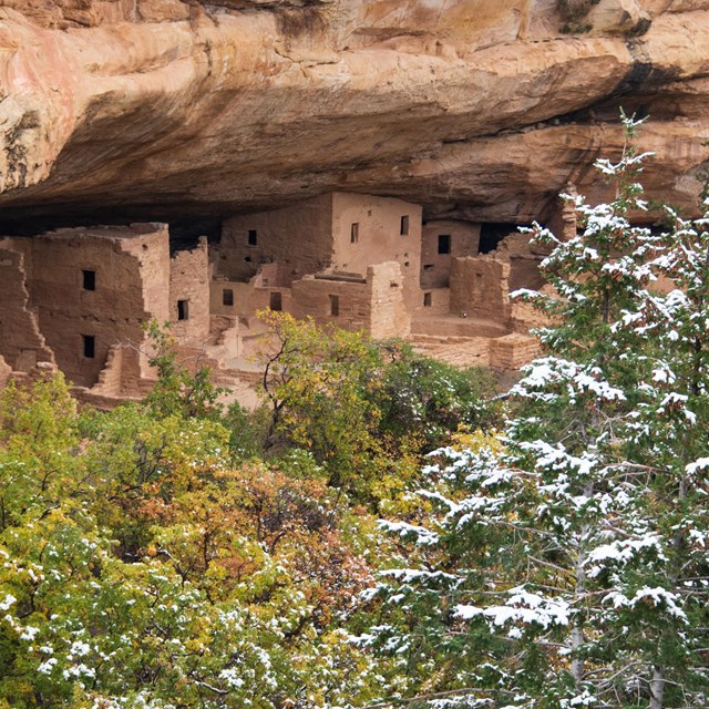 A stone masonry village under a sandstone alcove and behind snow-covered firs