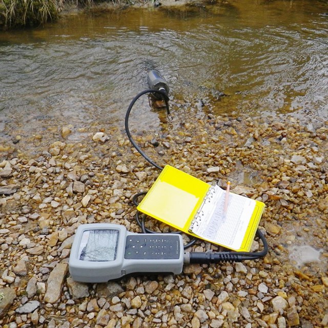 Device recording water chemistry at the edge of a stream beside a data sheet and pencil