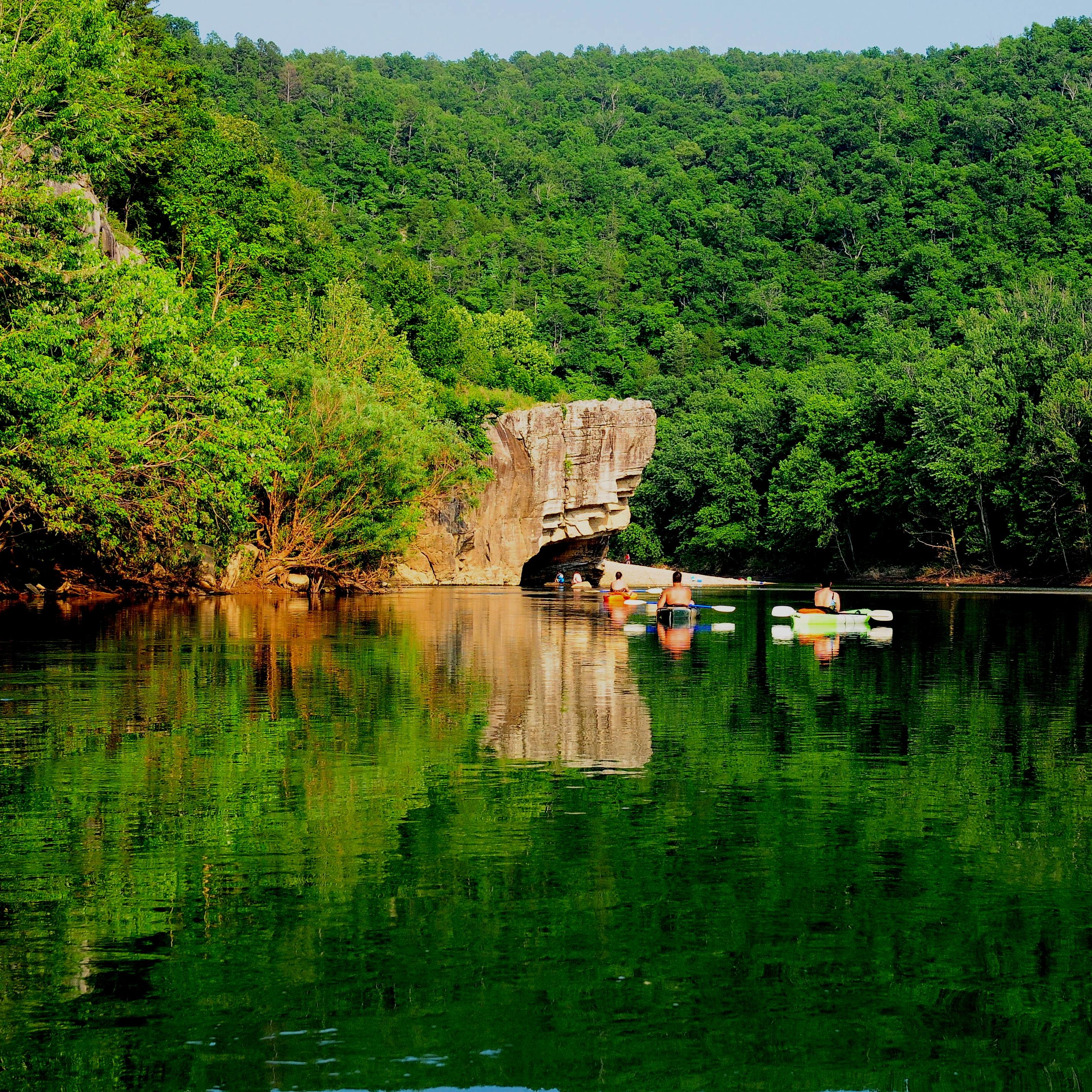 Kayakers float towards a rock on a river under lush green forested hills. 