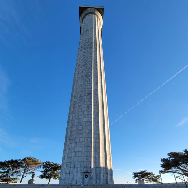 View of a large cylindrical brick column rising into a blue sky from steps below. 