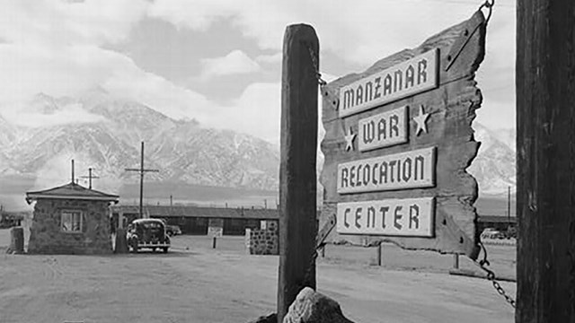 Black and white image of Manzanar sign with buildings in the background.