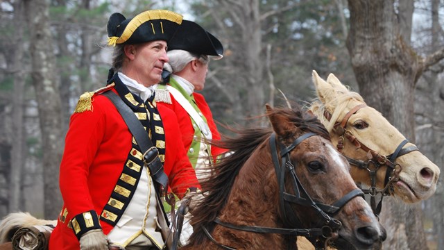 A British officer mounted on horseback with a scarlet red coat. The officer rides through trees