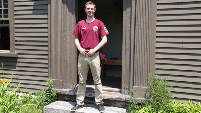 An intern in a red shirt stands in the doorway of a historic wooden house