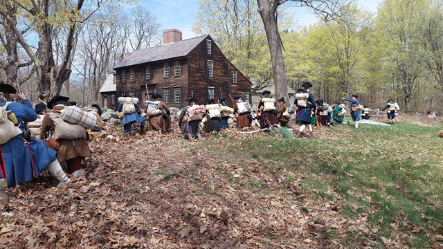 A group of colonial militia soldiers hide behind a stone wall in front of a wooden tavern house.