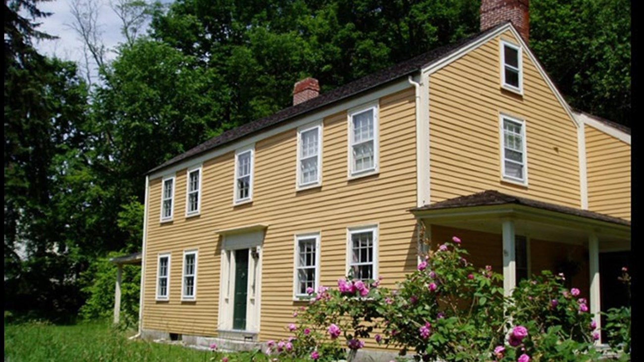 A yellow two and a half story historic house. A stone wall with flowering bushes runs in front