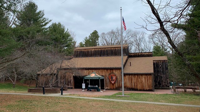 A large wooden building with tall windows is surrounded by tall trees. A tent and flag are at front.