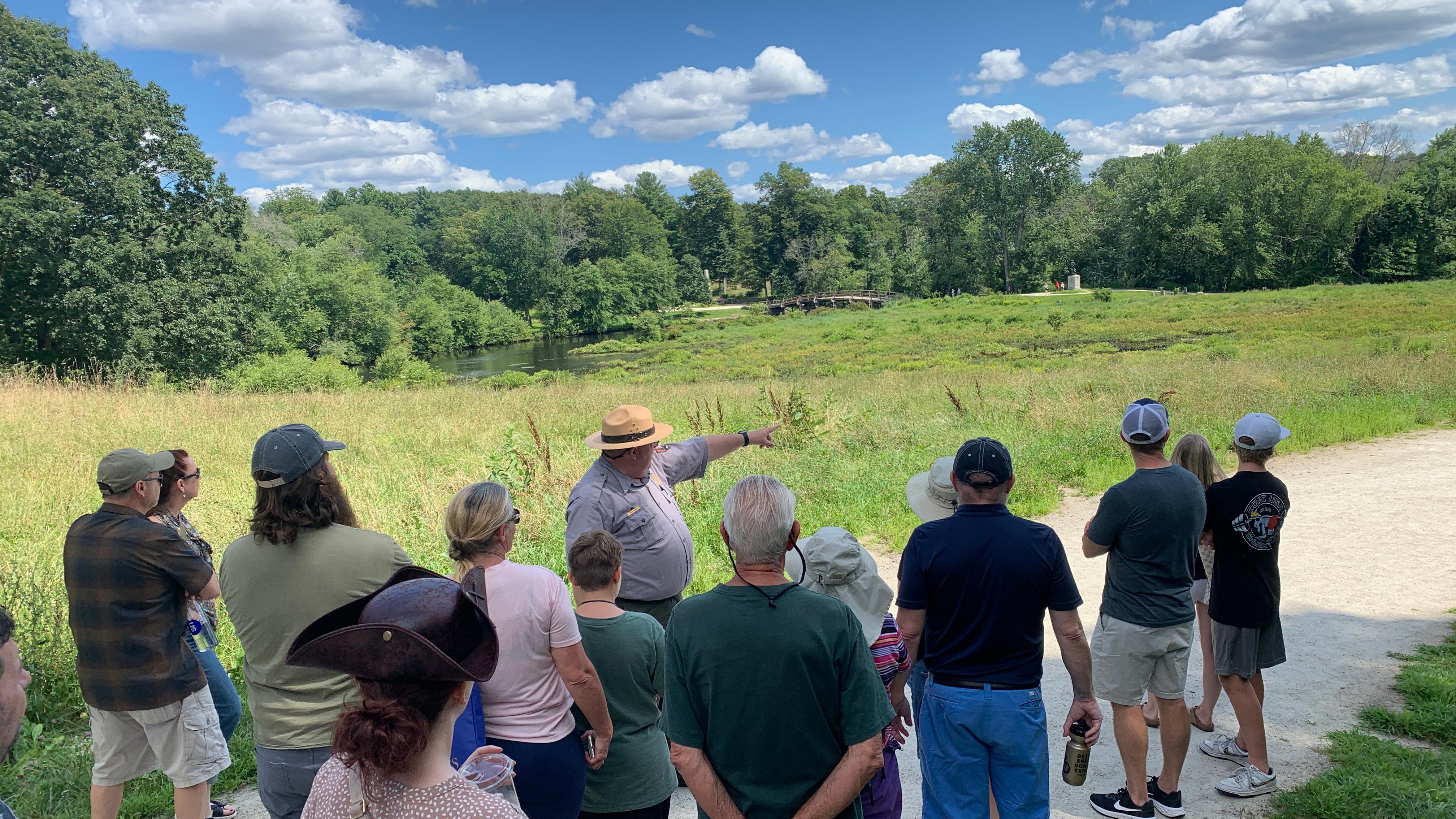 A park ranger in flat hat stands in front of a visitor group. The ranger points at a wide landscape.