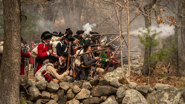 Militia soldiers crouch behind a stone wall loading and firing muskets while surrounded by smoke.