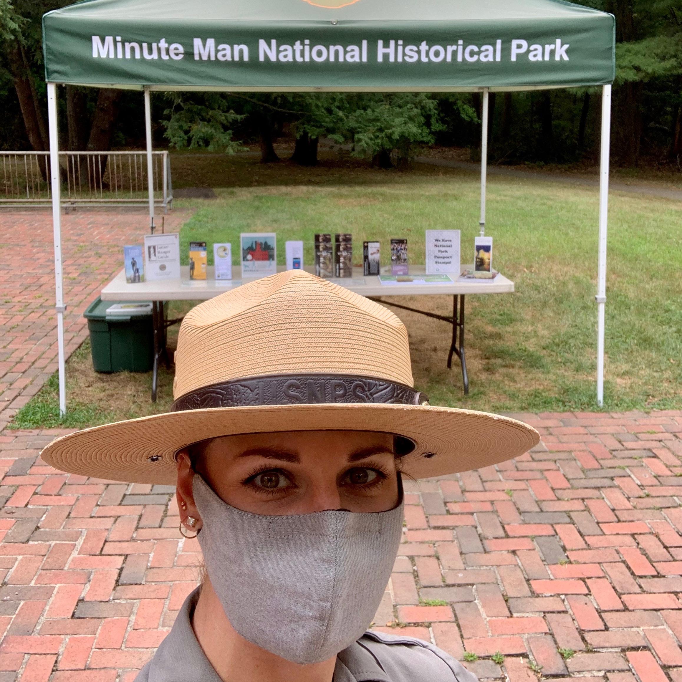 Female ranger wearing a mask that covers her nose and mouth stands in front of an outdoor info table