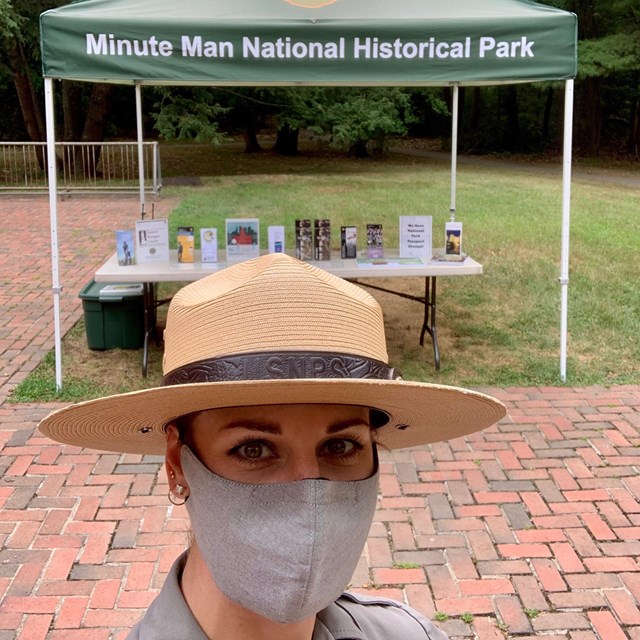 Female ranger wearing a mask that covers her nose and mouth stands in front of an outdoor info table