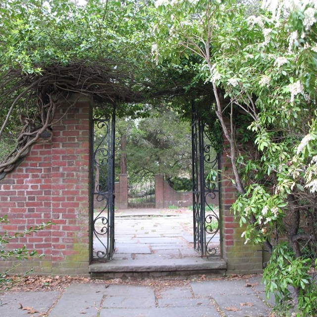 Gateway of red brick and iron stands open and surrounded by bushes