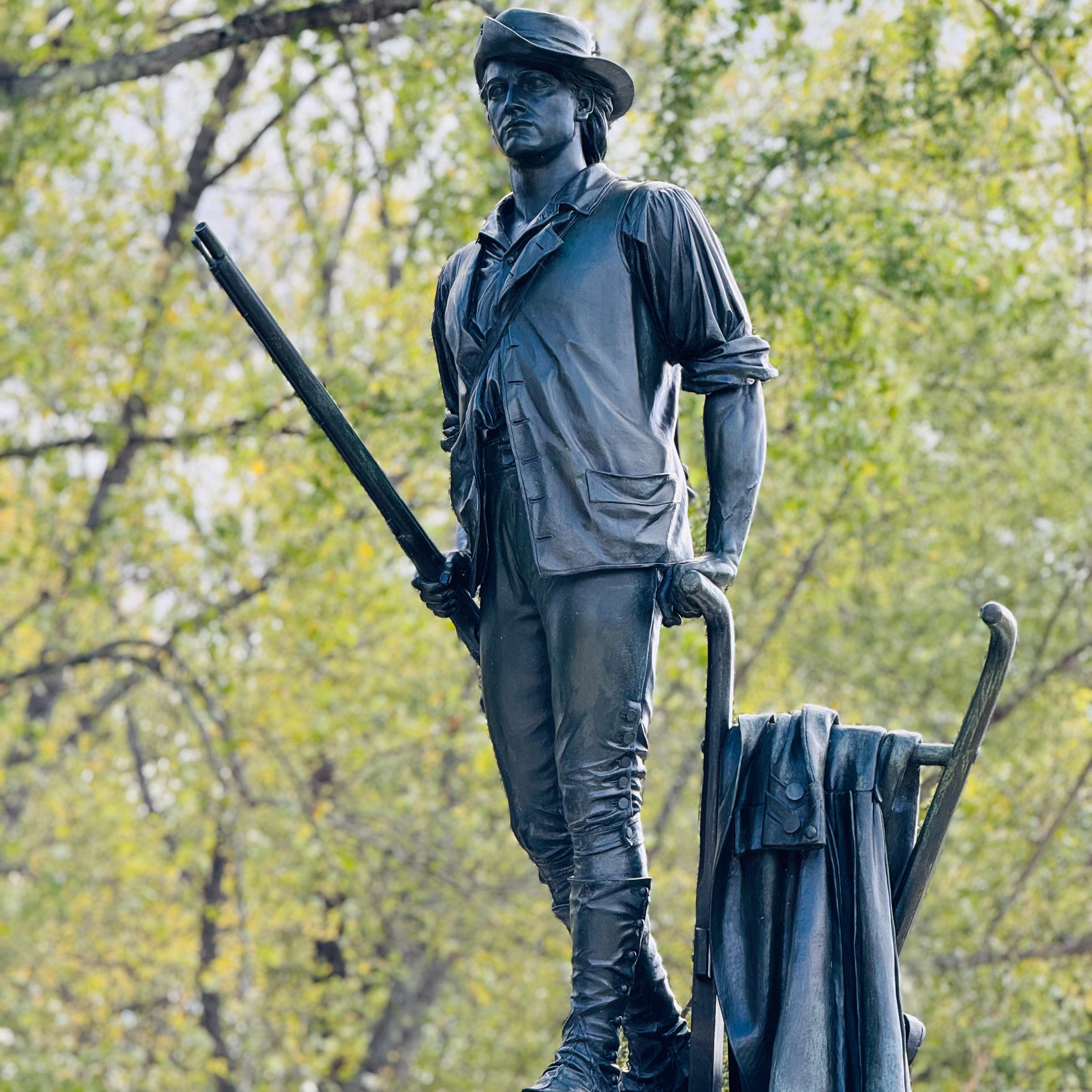 A bronze statue of a minute man sits atop a granite base. The man is walking and carrying a musket