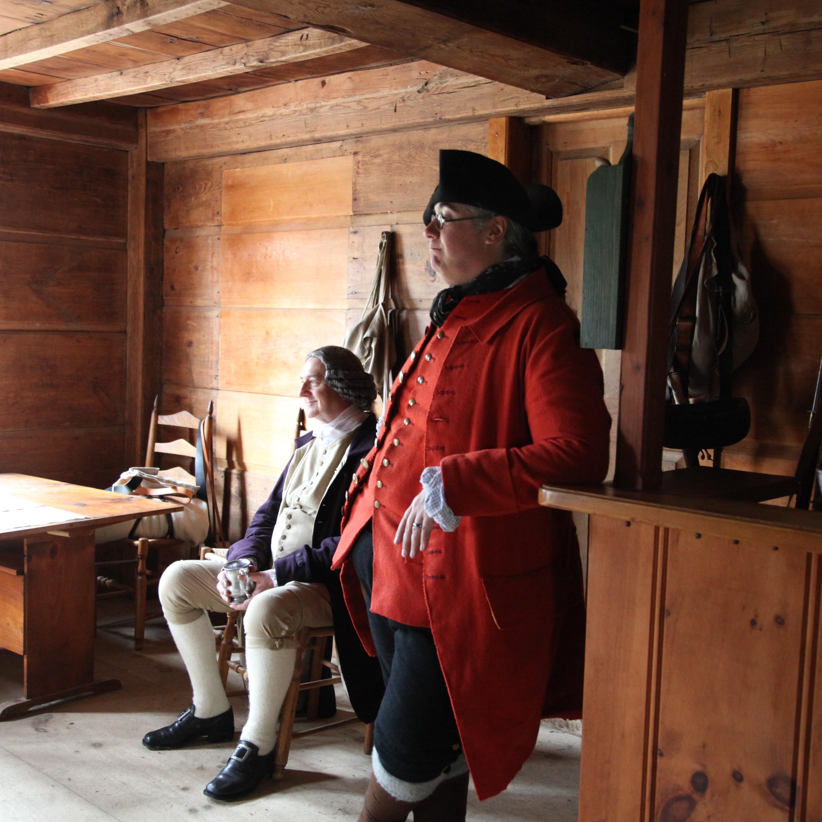 A man dressed in 18th century clothing leans against the bar next to a man sitting at a table