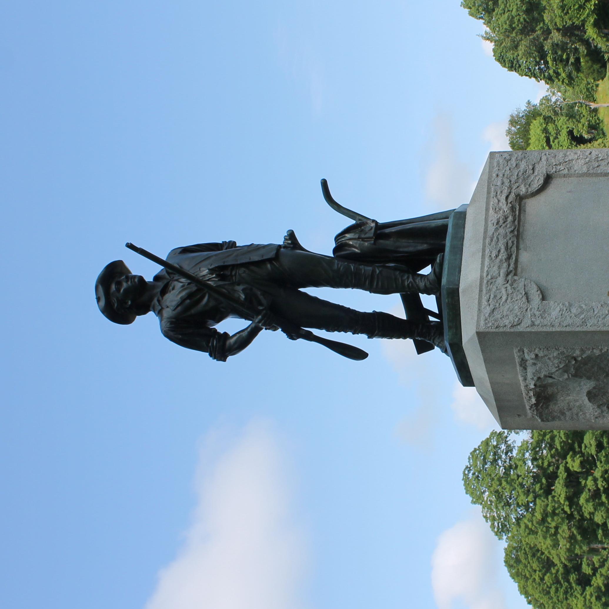 A bronze statue of a minute man sits atop a granite base. The man is walking and carrying a musket