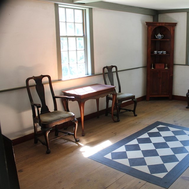 A room in a historic house with a wooden floor, checkerboard floor cloth, side chairs and a table