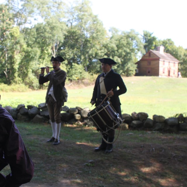 A colonial fifer and drummer stand in front of a stone wall. A colonial house is in the background.