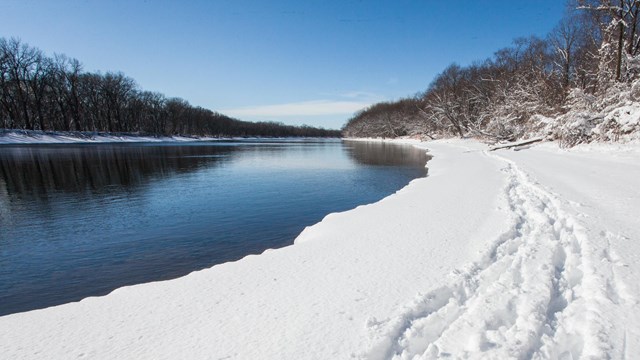 Tracks lead through the snow along a curved riverbank. The river reflects the sky and trees.
