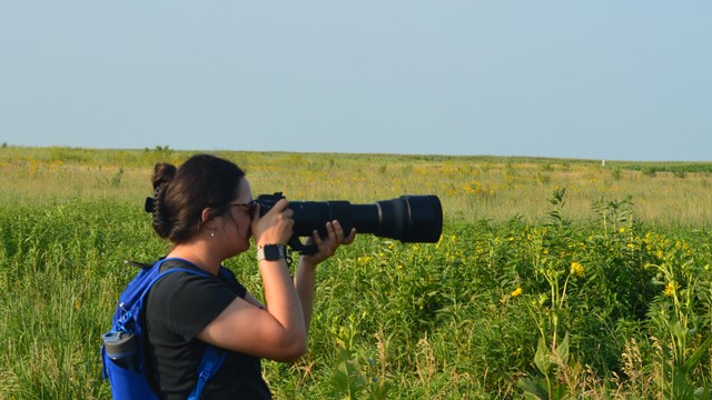 A female holding a large black camera to her face while standing in a field of tall green grass.