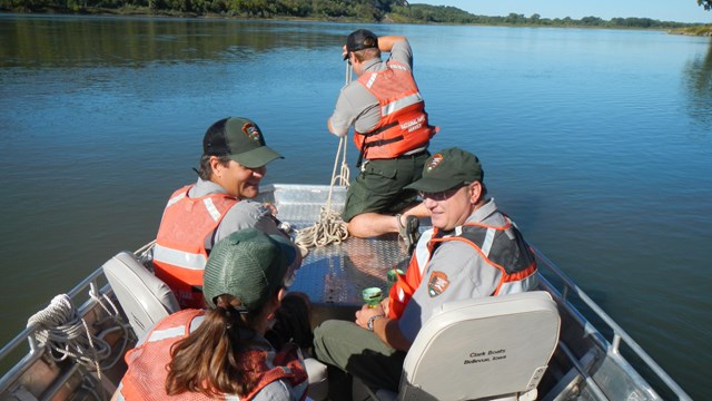 Four park rangers sit in a metal boat on the river discussing and conducting scientific research.
