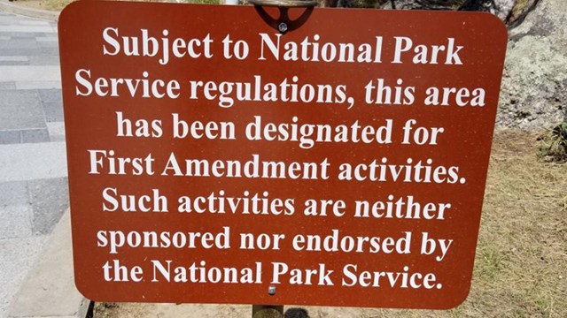 A park sign identifies the area as a first amendment area for activities and demonstrations.
