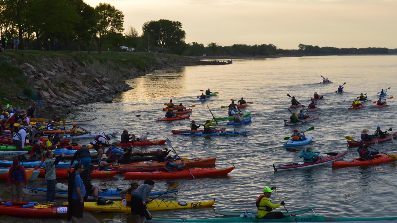 Numerous kayaks launch into a river from a sandy bank.