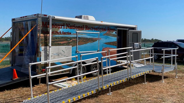 A large trailer with a long metal ramp attached is parked in a grassy area