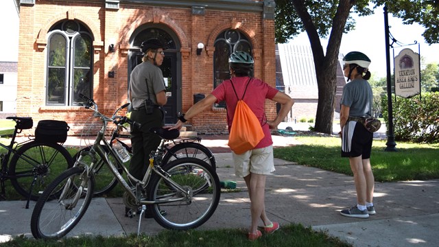 A park ranger on bike talks to two visitors in front of a historic brick building